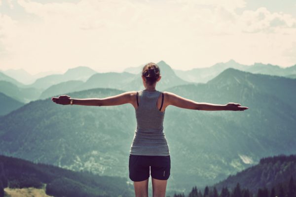 Woman celebrating nature and reaching the summit of a high mountain as she stands with her back to the camera and arms extended looking out over mountain ranges and valleys in a panoramic landscape